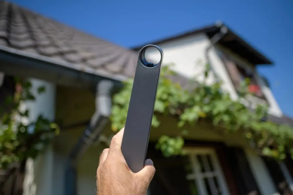 A hand holding a long Starlink wall mount for the antenna, with a house and clear blue sky in the background, showing preparation for installation.